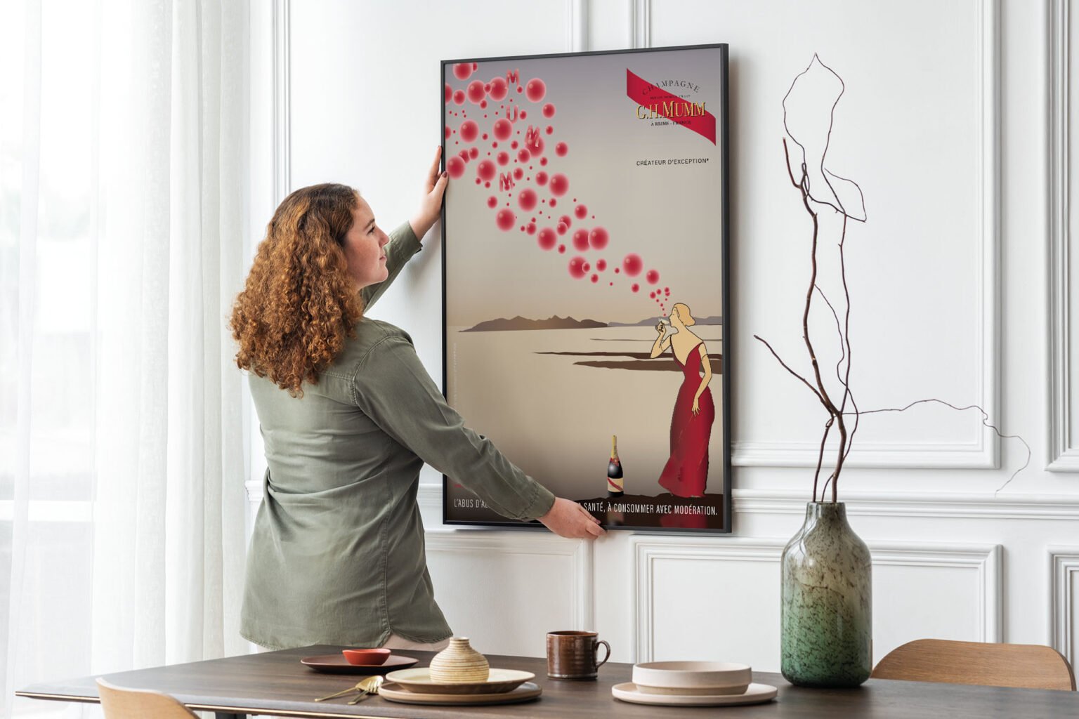 Une femme accroche au mur de son appartement une affiche encadrée. Il s'agit de l'affiche champagne Mumm qui représente une jeune femme blonde vue de profil, en robe rouge, une coupe de champagne à la main qu'elle porte à ses lèvres. Des bulles rouges de toutes tailles s'échappent de sa coupe. Derrière elle un paysage de lacs et de lointaines montagnes.
