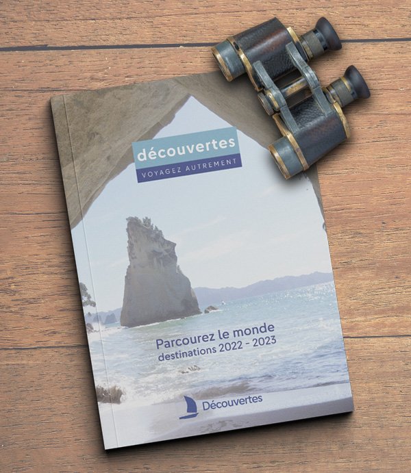 Brochure de l'Agence de Voyage Découvertes. La couverture présente un paysage de bord de mer avec de hauts rochers. La brochure est représentée sur du bois brut. Des jumelles anciennes sont posées par-dessus.