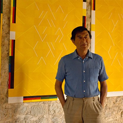 Photo du peintre Albert Ayme devant deux de ses toiles en hommage à Van Gogh. Ces toiles abstraites à dominante jaune, sans chassis, sont accrochées sur un mur brut.